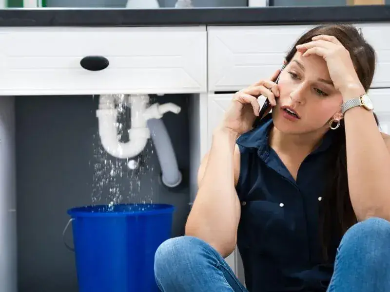 Homeowner calling for help beside a leaking pipe under a kitchen sink